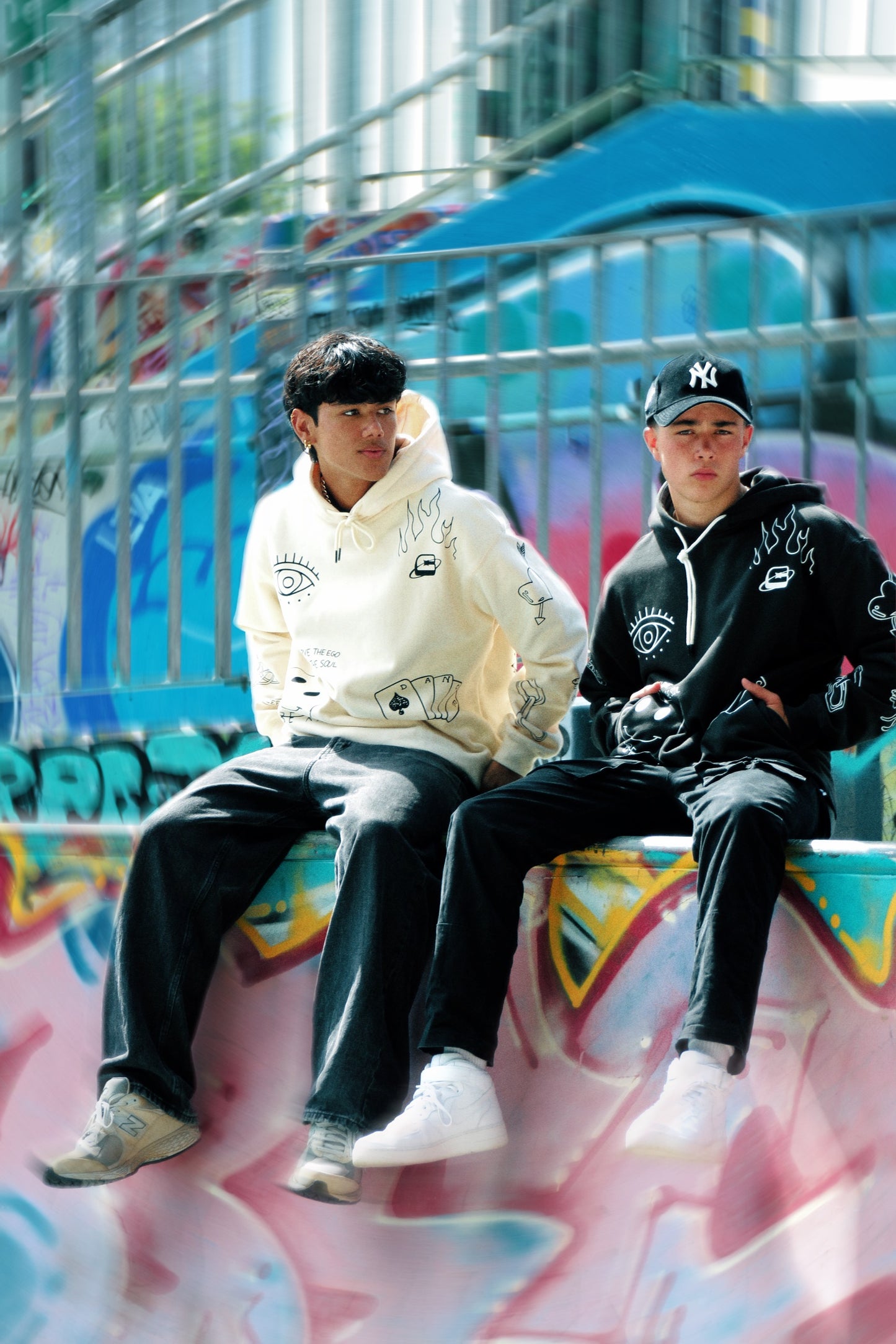 Two individuals wearing oversized black hoodies against a colourful graffiti background, showcasing casual streetwear style.