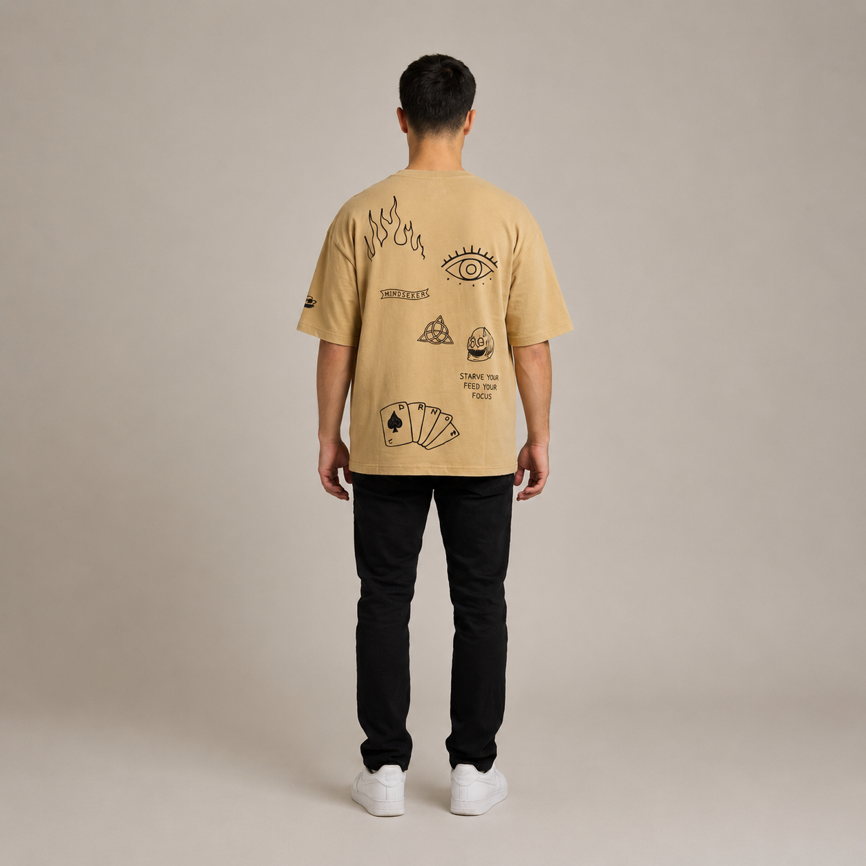 Tan oversized t-shirt with graphic prints of an eye, flames, and playing cards, styled with black pants and white shoes.