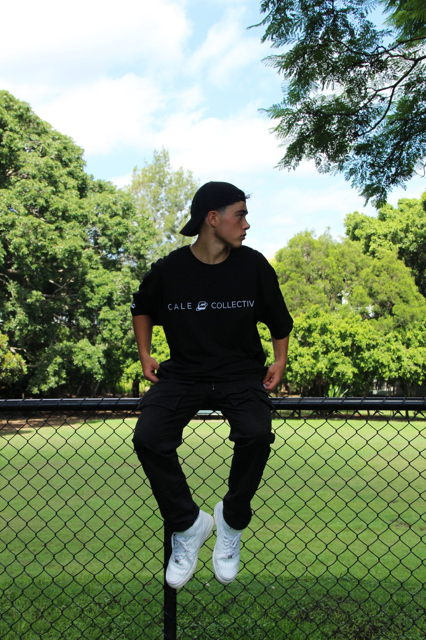Person wearing a black oversized graphic t-shirt with 'CALE COLLECTIVE' text, paired with black pants and white shoes in a green setting.