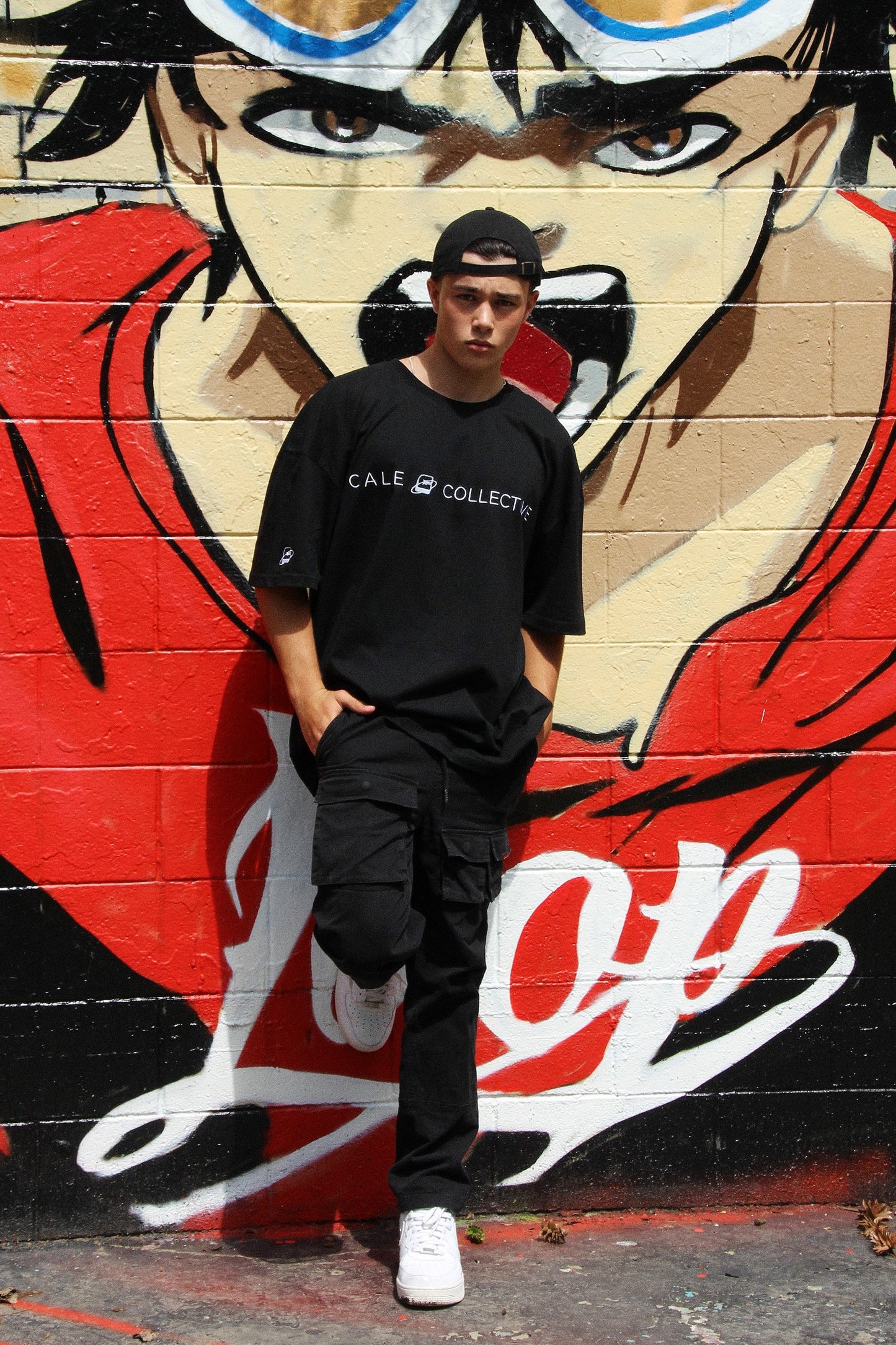 Black oversized graphic t-shirt worn by a model with dark pants and white sneakers against a colourful mural background.