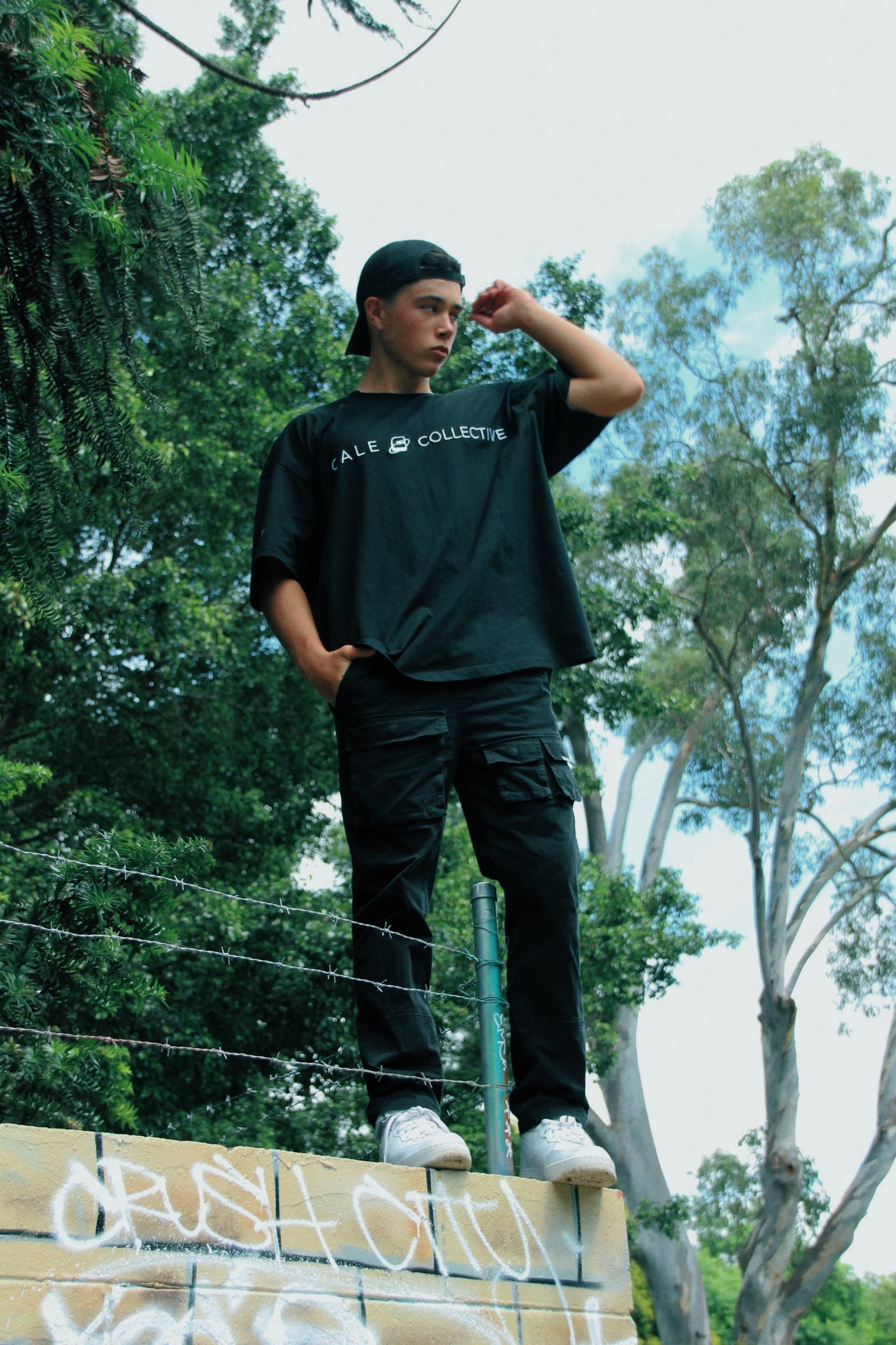 Black oversized graphic t-shirt with 'CALE COLLECTIVE' text, worn with black cargo pants and white sneakers against a graffiti backdrop.