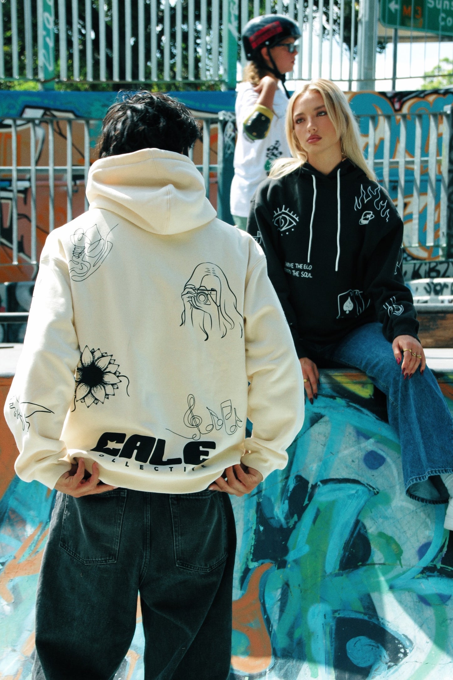 Two individuals in an urban skate park wearing black oversized hoodies with graphic designs on the back.