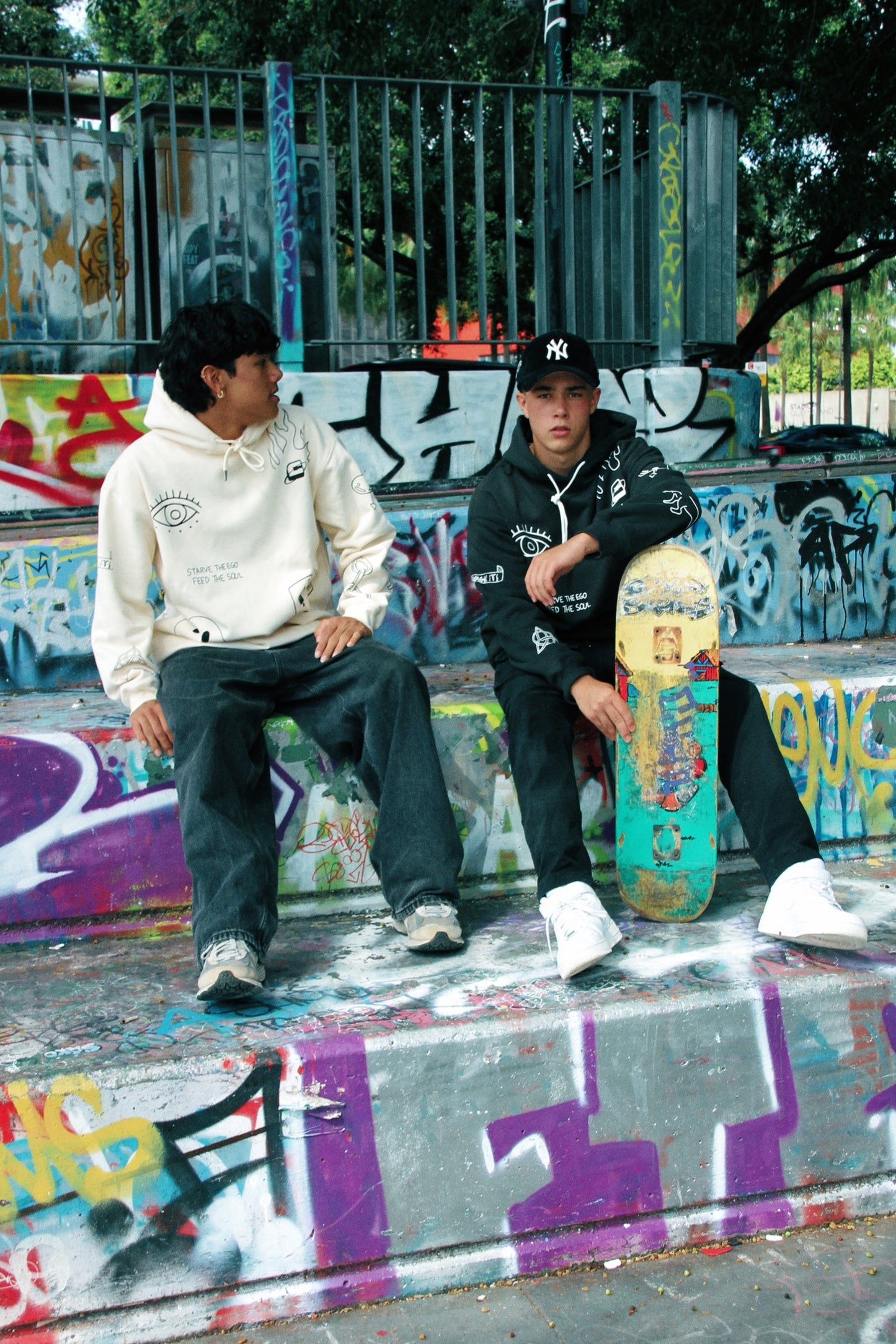 Two individuals sitting on graffiti-covered steps, one wearing a black oversized hoodie with graphic detailing.