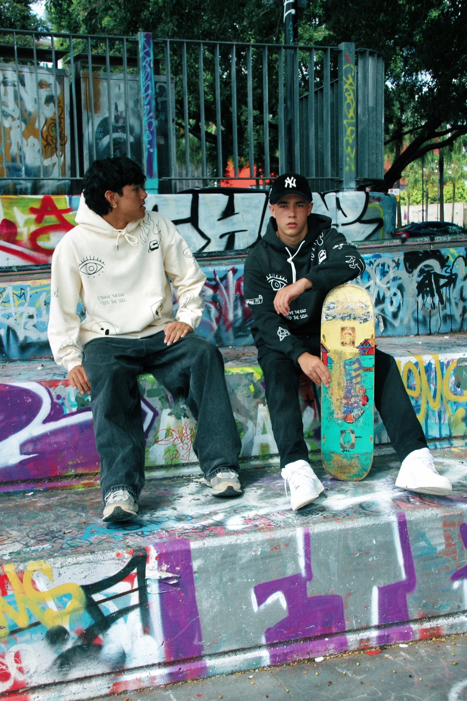 Two individuals sitting on graffiti-covered steps, one wearing a black oversized hoodie with graphic detailing.